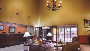 A spacious lobby with a brown leather sofa, two armchairs, a coffee table, a chandelier, a clock, and a reception desk. - La Quinta Inn & Suites by Wyndham Alice (Alice)