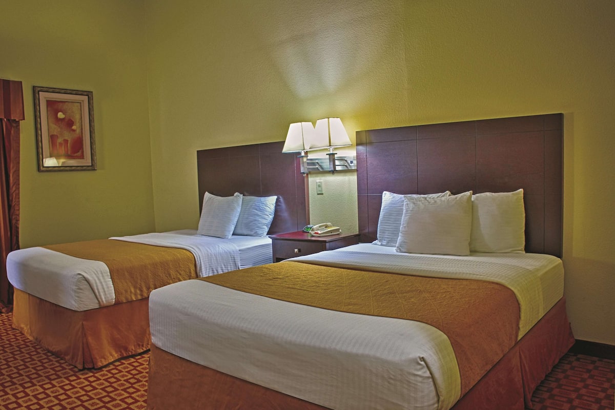 A hotel room with two beds, each with a white comforter and a brown bed skirt. There is a nightstand between the beds with a lamp and a telephone.