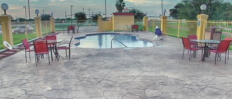 Un área de piscina con suelo de piedra hexagonal, sillas y mesas rojas, rodeada por una valla amarilla con farolas.