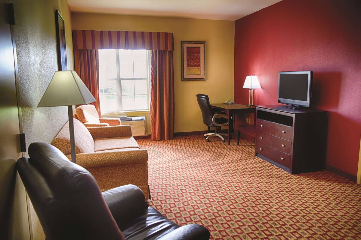 A hotel room with a red carpet, a sofa, a chair, a desk with a lamp, and a television.