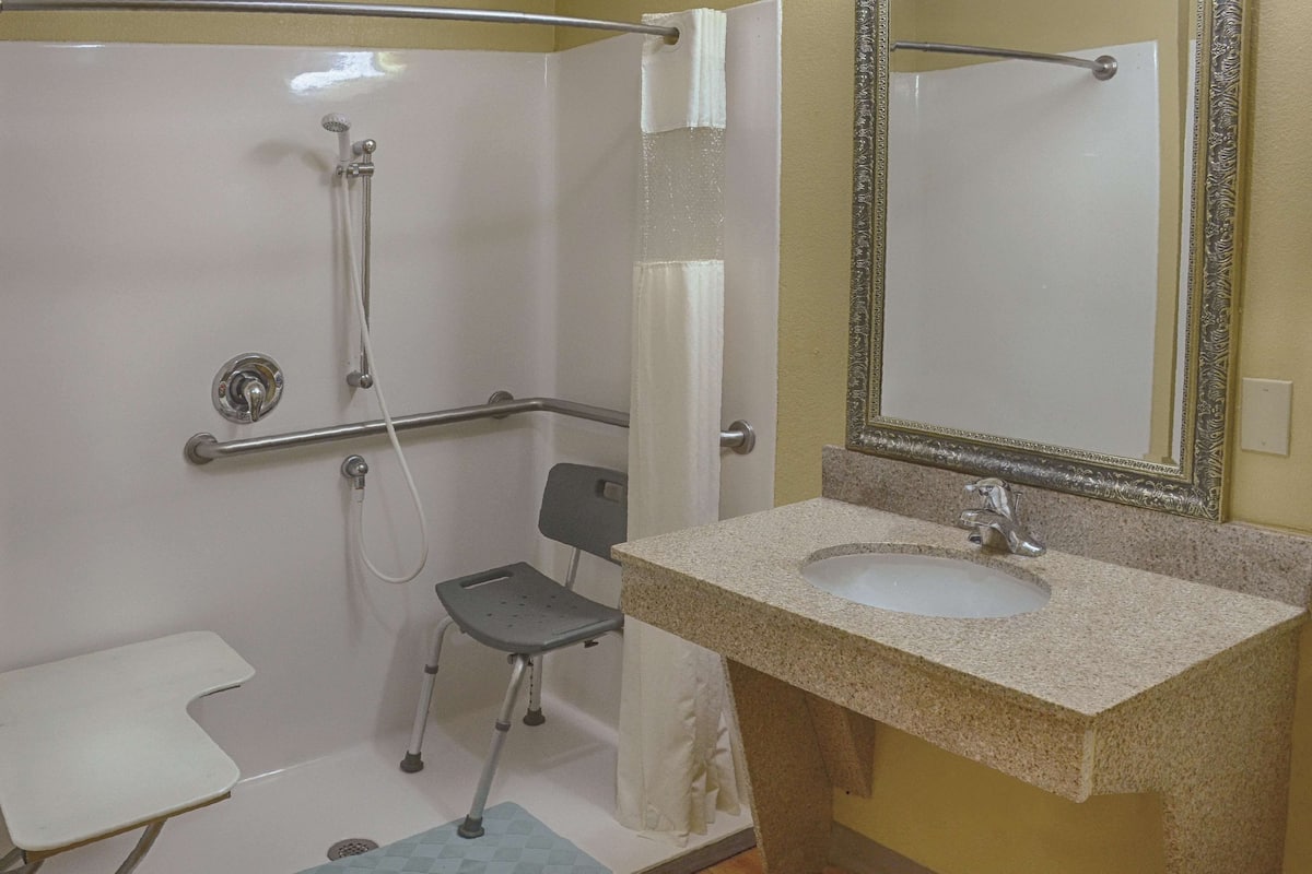 A bathroom with a shower, grab bar, wheelchair-accessible chair, sink, and toilet.