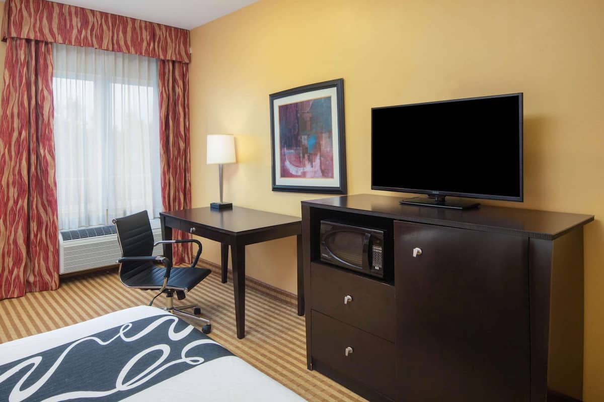 Habitación de hotel con cama, escritorio con silla, televisor, microondas, un cuadro enmarcado en la pared y ventana con cortinas.