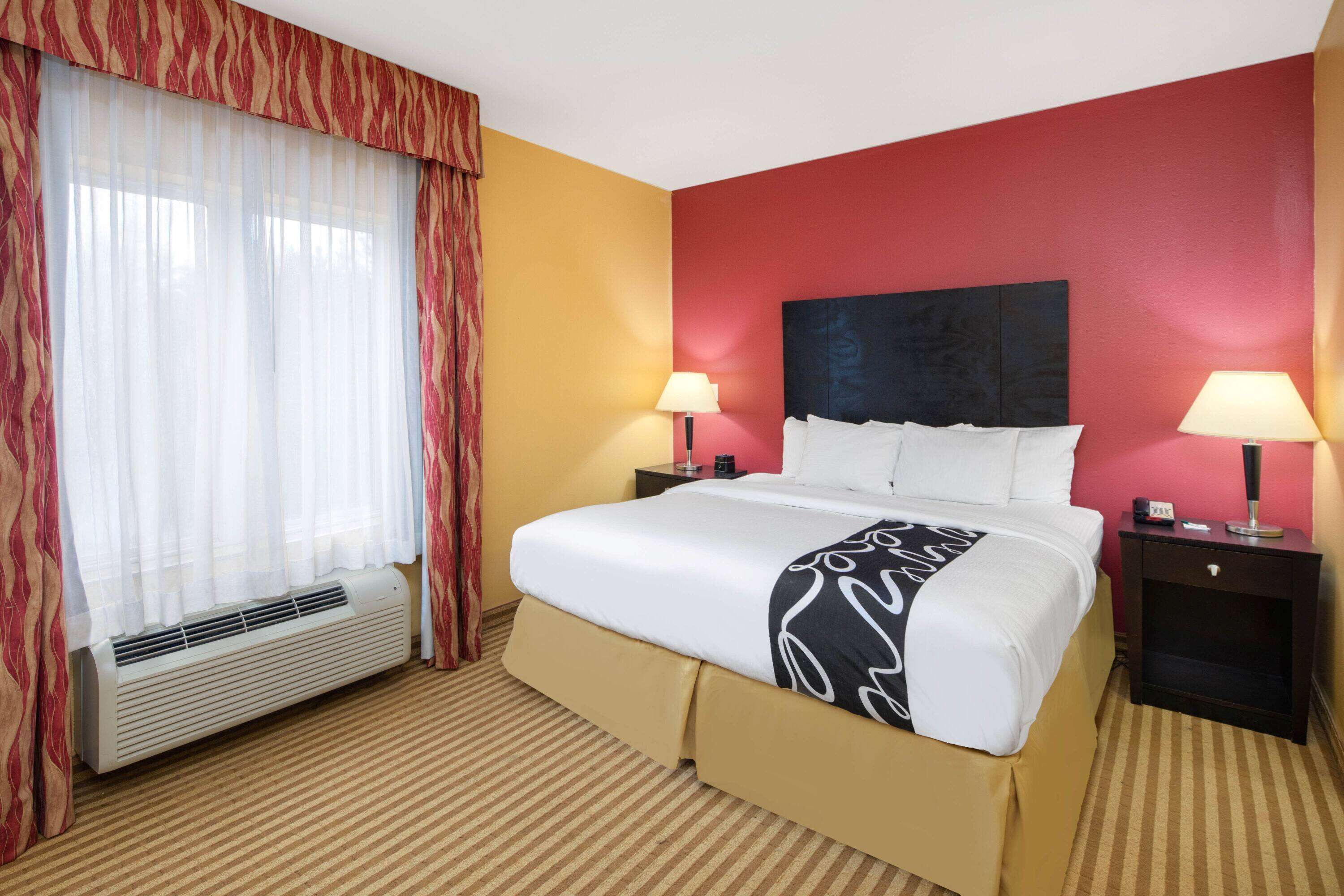A hotel room with a large bed, two bedside tables, a window with curtains, and a red accent wall.