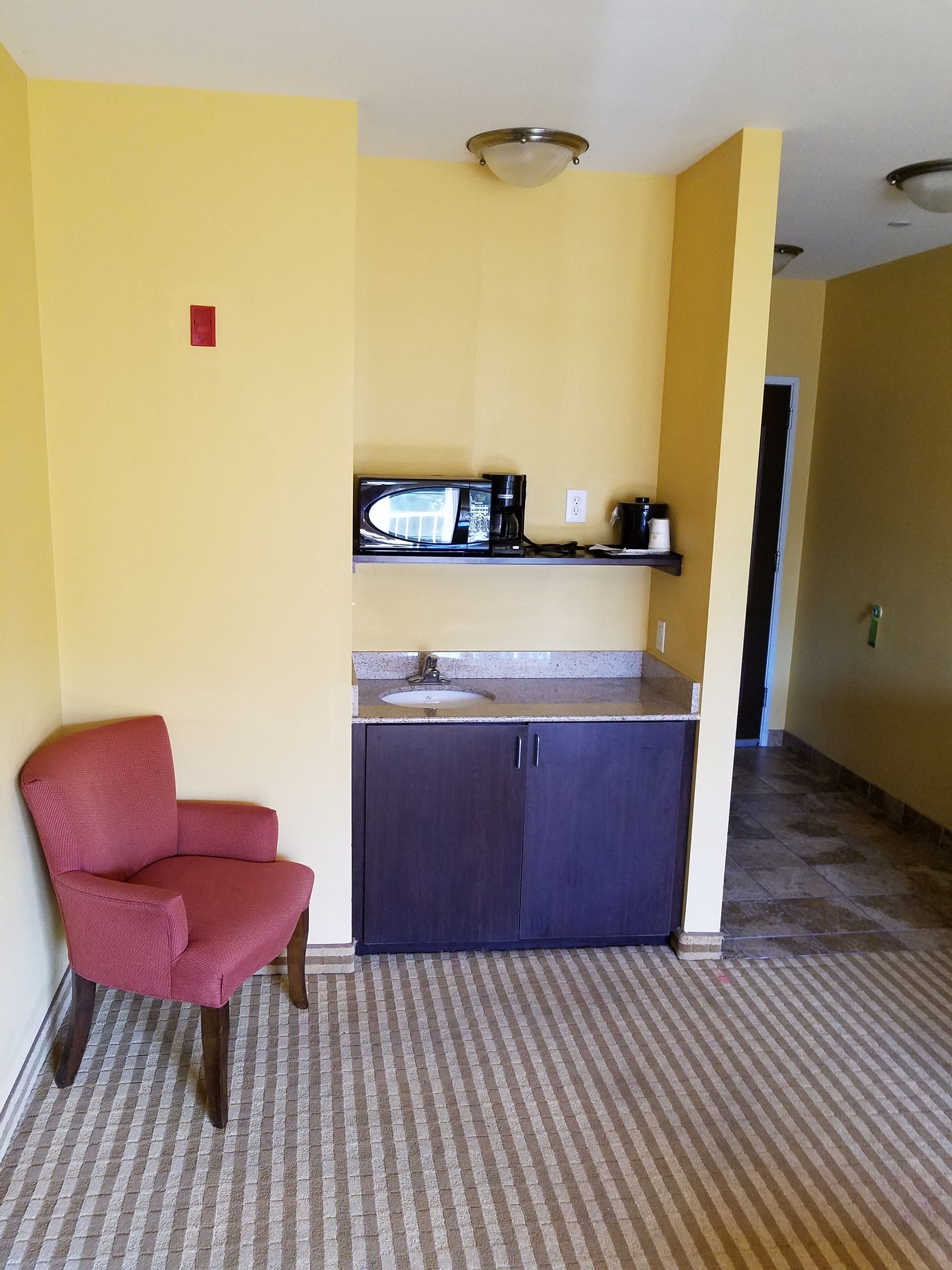 A small kitchenette with a microwave, coffee maker, and sink. There is a red chair and a patterned carpet.