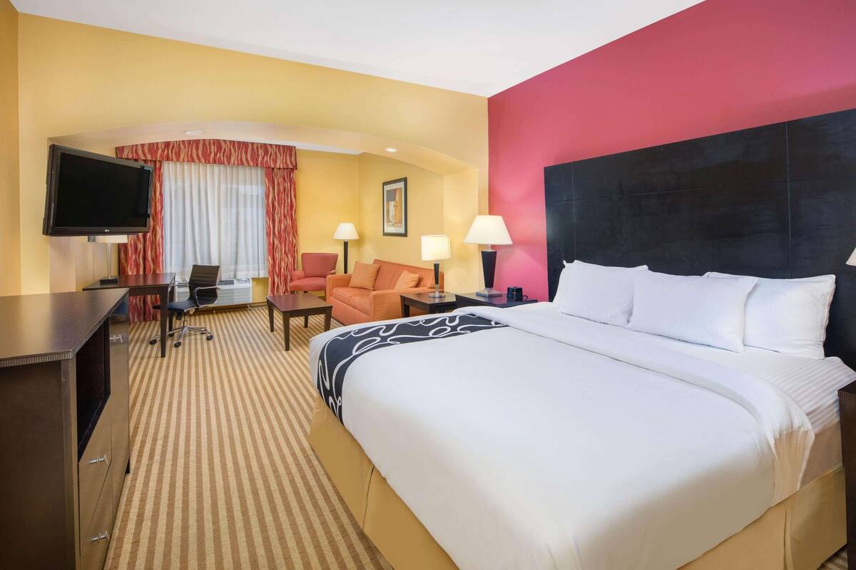 Habitación de hotel con una cama grande, un escritorio con televisor, una silla, dos lámparas y una ventana con cortinas.