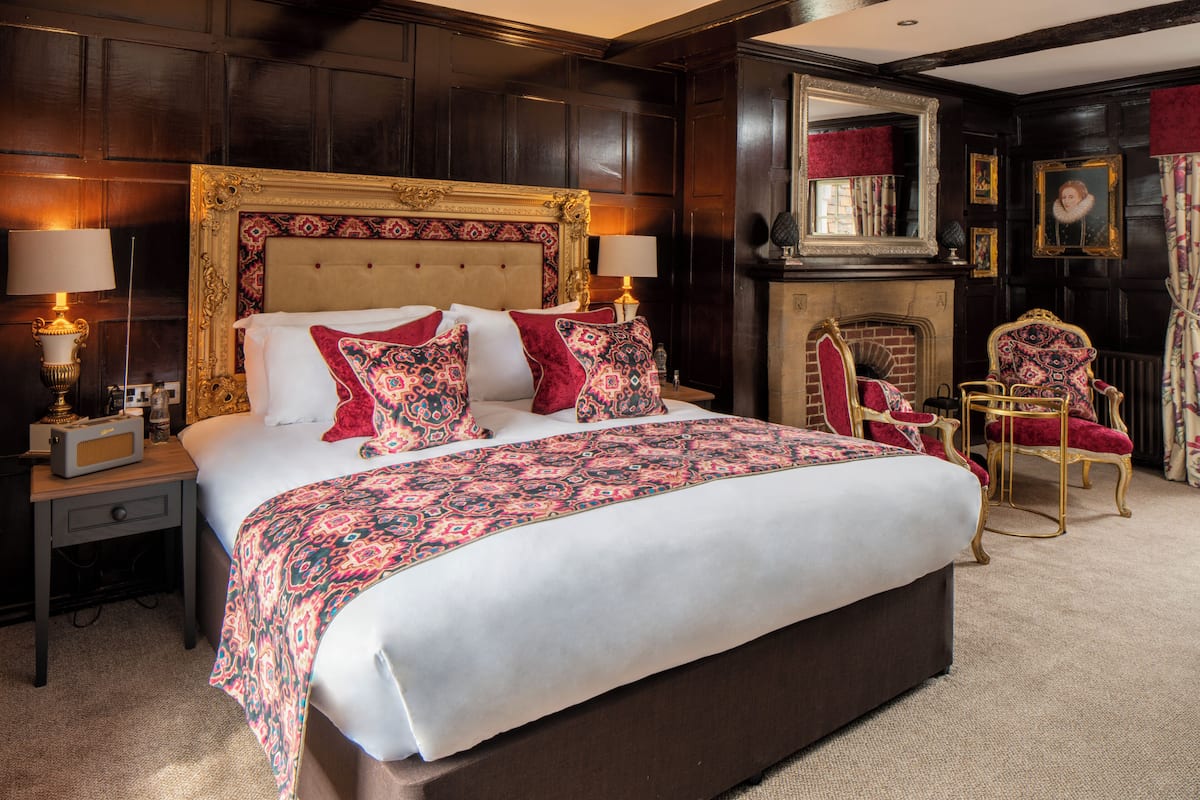 A large bed with a patterned bedspread, two bedside tables with lamps, a fireplace, and ornate chairs.