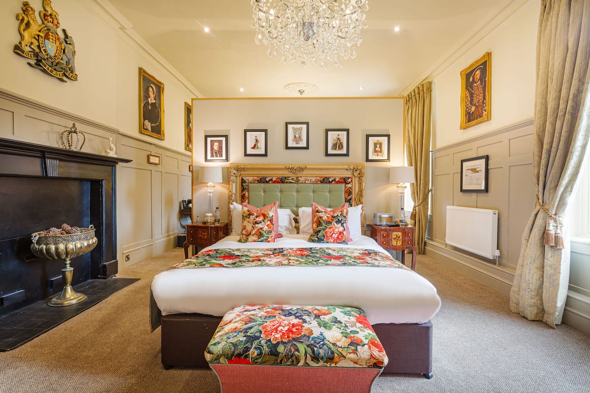 A large bed with a floral bedspread, a chandelier, framed pictures on the wall, and a fireplace.