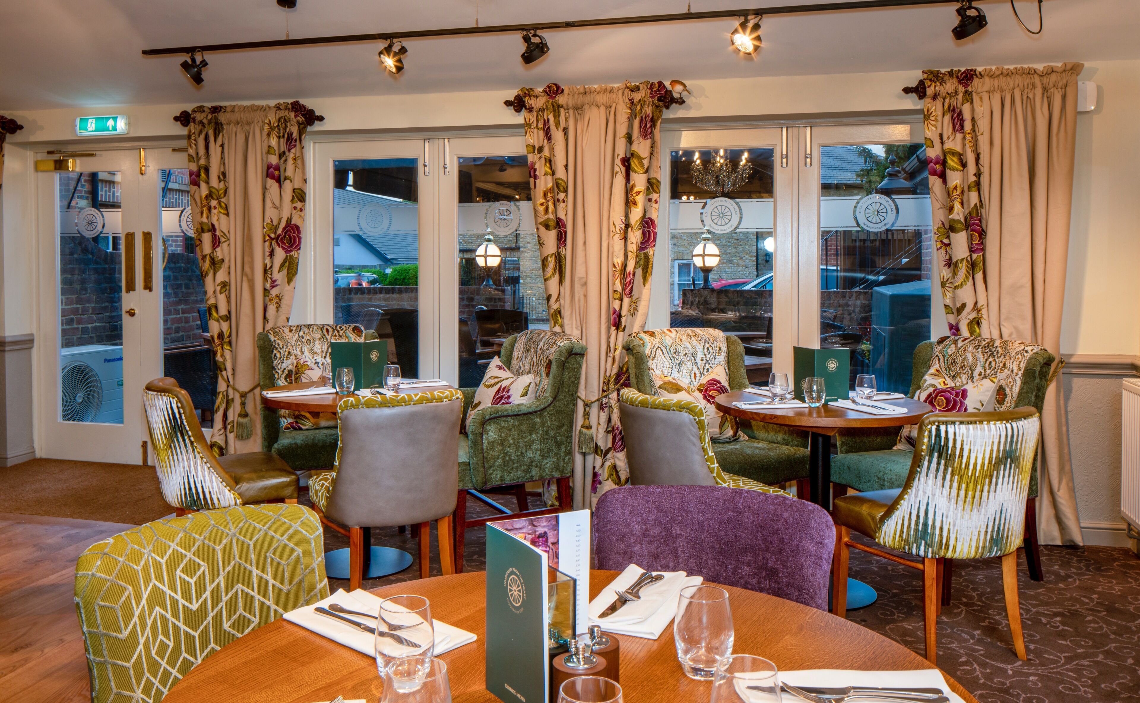 A restaurant with a cozy interior, featuring multiple dining tables and chairs, large windows with curtains, and a visible exit sign.