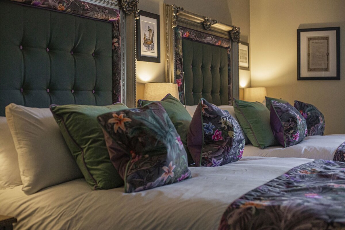 A hotel room with a tufted headboard, floral-patterned pillows, and framed artwork on the walls.