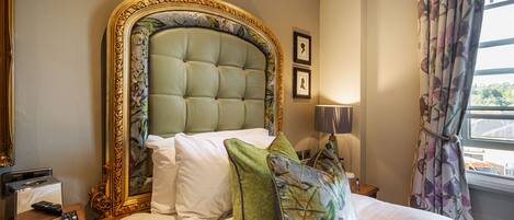 Habitación de hotel con una cama grande, cabecero con motivos florales, mesita de noche con lámpara y una ventana con cortinas.