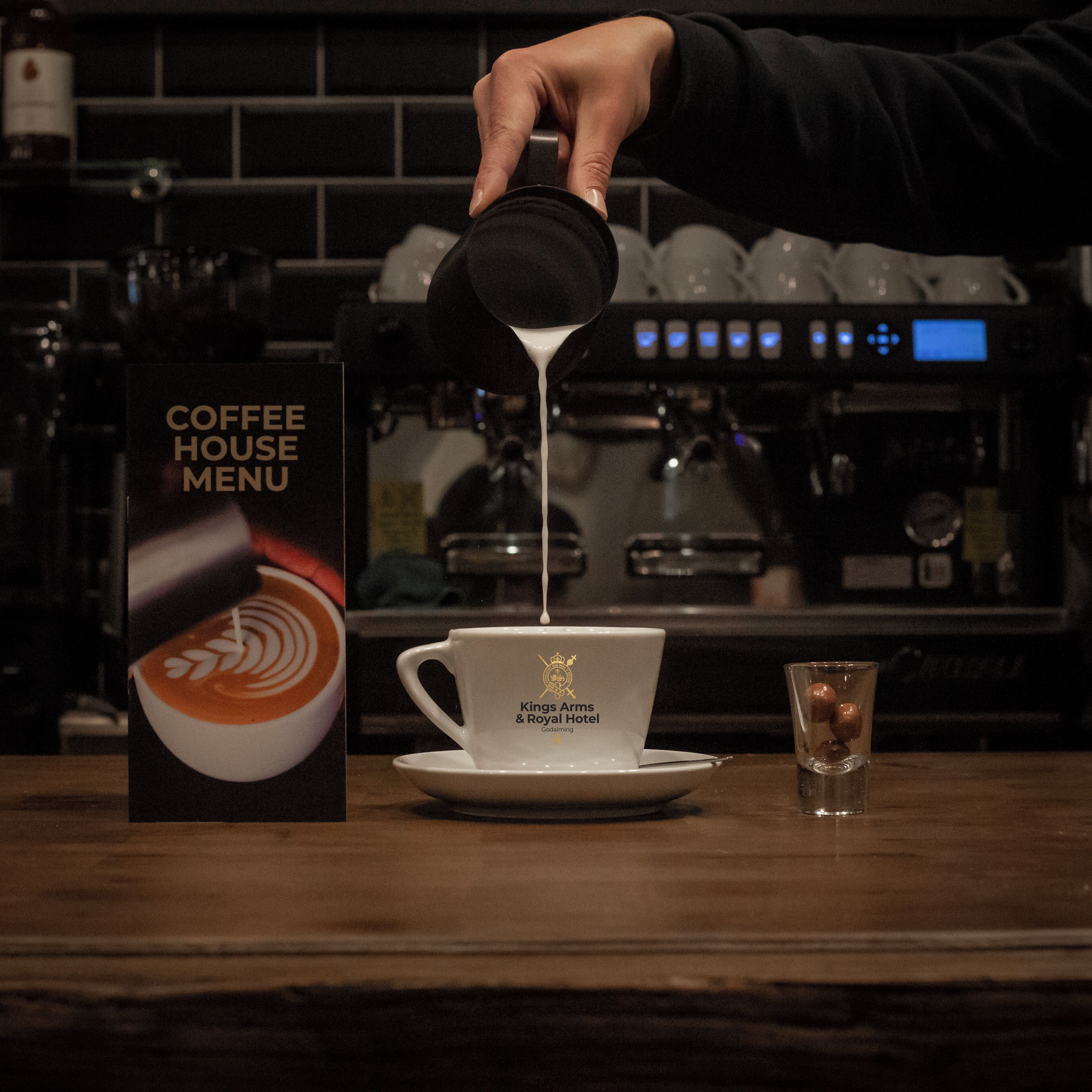 A hand pouring coffee into a cup with a logo, a menu stand, and a coffee machine in the background.