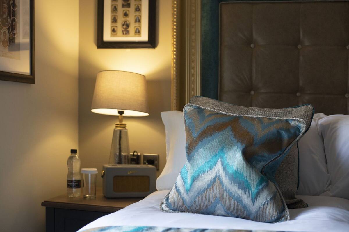 A hotel room with a bed, a nightstand with a lamp, a radio, and a framed photo on the wall.