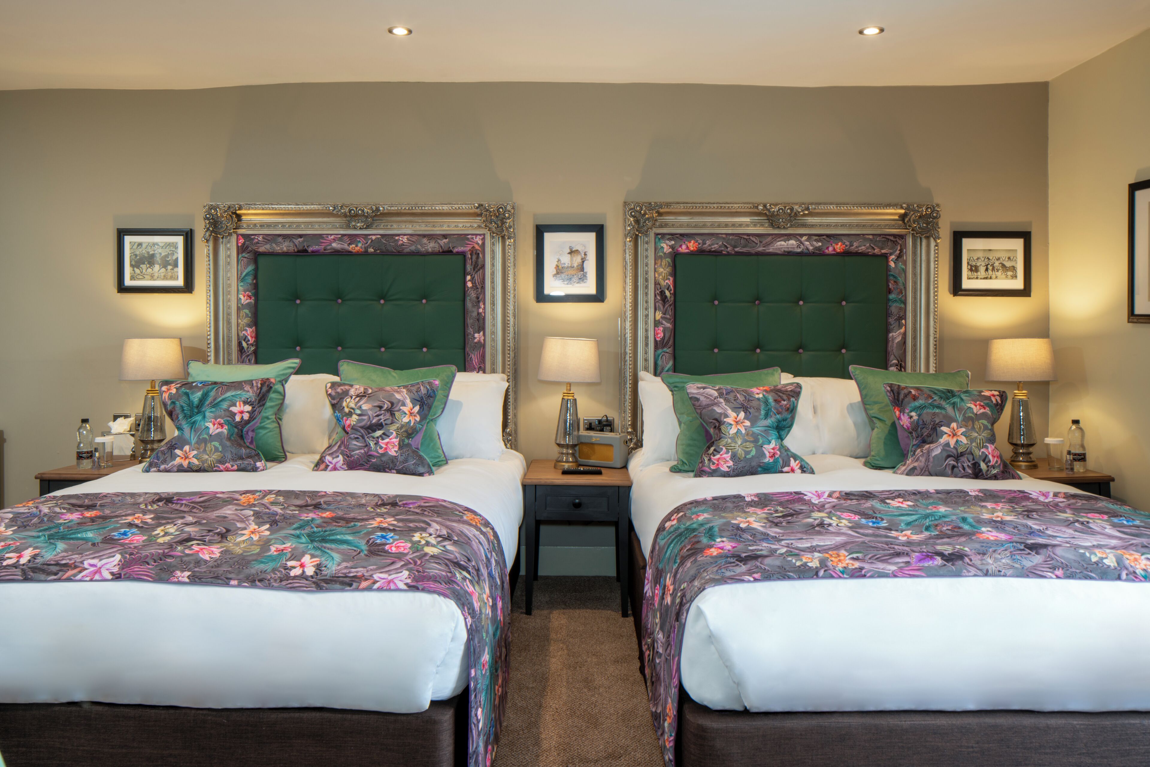 A double room with two beds, each with a green upholstered headboard, flanked by nightstands with lamps and framed pictures on the walls.