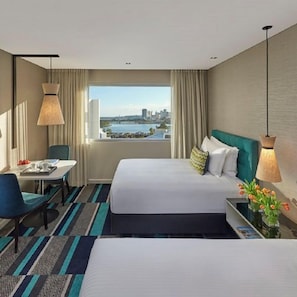 Habitación de hotel con dos camas, un escritorio, una silla, vistas a la ciudad y un ventanal con cortinas.