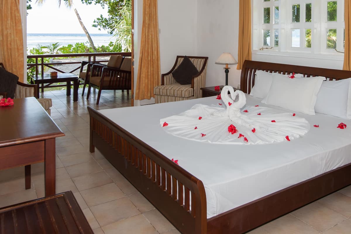 Habitación de hotel con una cama decorada con un cisne y pétalos de rosa, cabecero de madera, mesita de noche con lámpara, una silla y vista al océano a través de grandes ventanales.