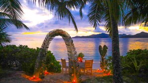 A beach setting with a table and chairs under a palm archway, overlooking a calm sea and distant mountains.