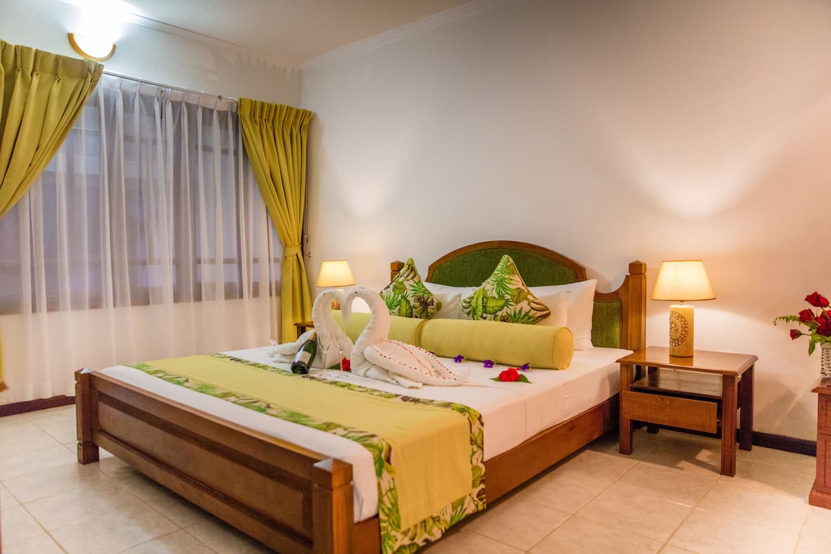 Una cama bien hecha con una colcha verde, dos almohadas, una lámpara de noche y una mesita de noche con un florero de flores.