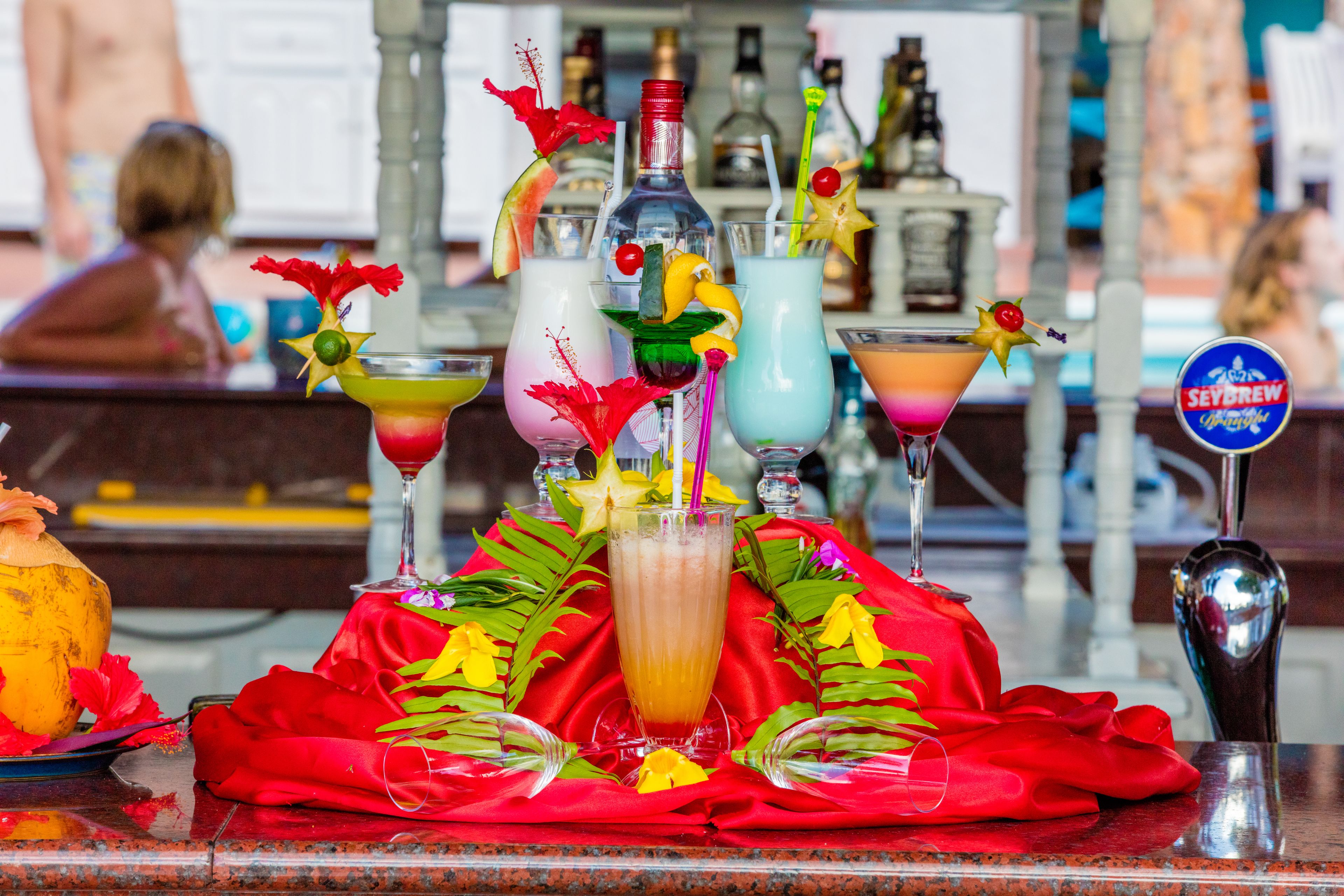 Zona de bar con varios cócteles y botellas, un paño rojo y un letrero con la marca 'SEYBREW'.
