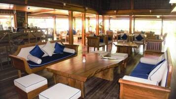 A wooden lounge area with cushioned seating and tables, large windows, and a view of the outdoors.