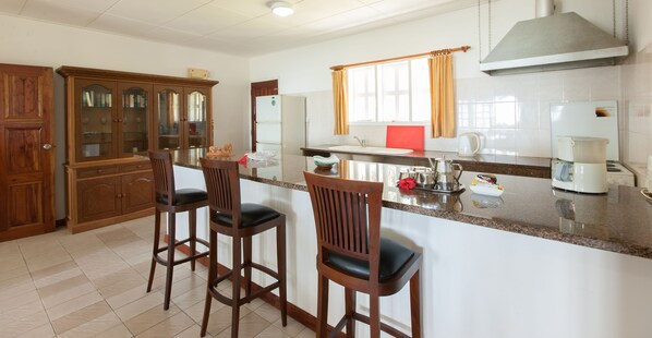A kitchen with a marble countertop, wooden chairs, a refrigerator, and a window with curtains. - La Digue Island Lodge (La Digue)