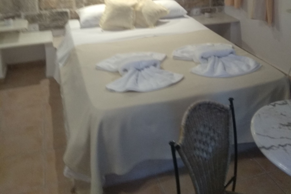 A bedroom with a stone wall, a bed with white linens, a nightstand, a chair, and a small table.