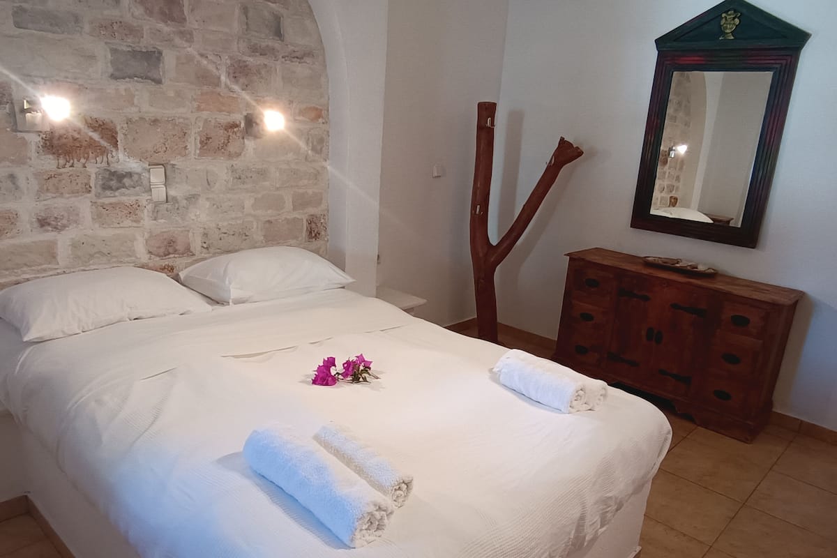 Una cama bien tendida con sábanas blancas y dos toallas enrolladas, un tocador de madera con espejo, y una pared de piedra con iluminación empotrada.