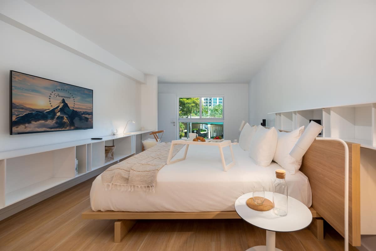 A modern bedroom with a bed, a bedside table, a TV mounted on the wall, and a window with a view of greenery.