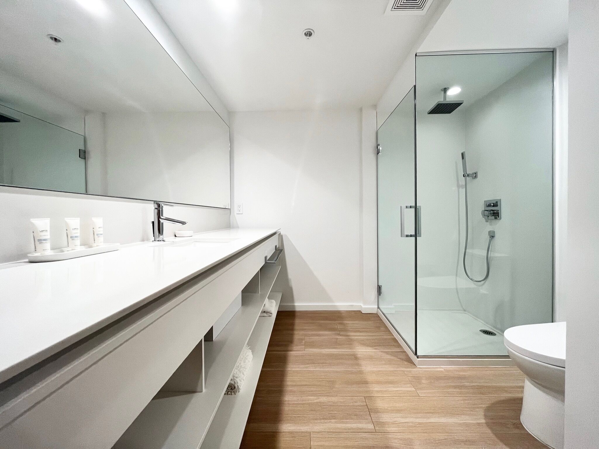 Un baño moderno con un espejo grande, una encimera blanca y una ducha con mampara de vidrio.