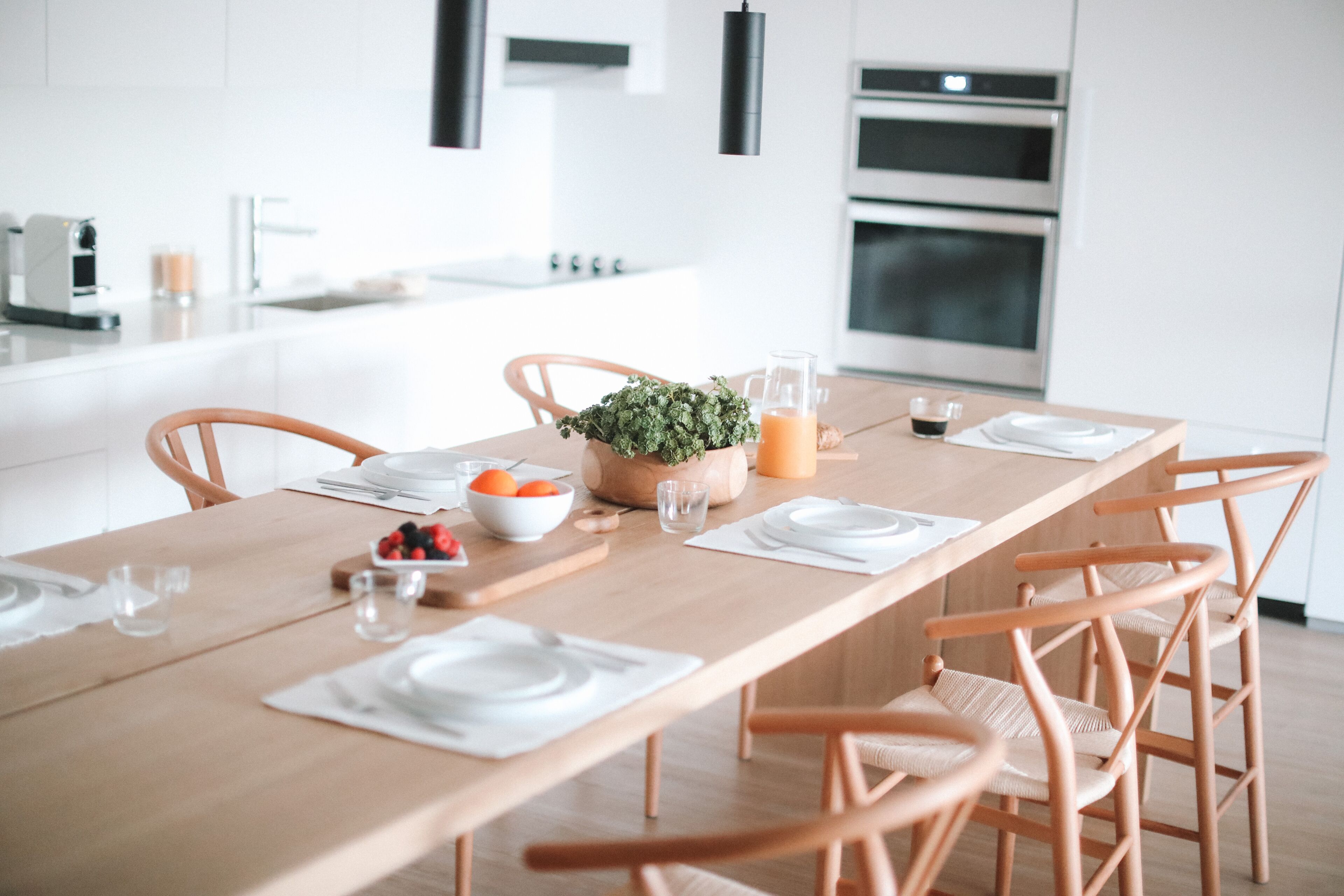 A modern kitchen with a wooden dining table set for four, featuring white plates, glasses, and a bowl of fruit.