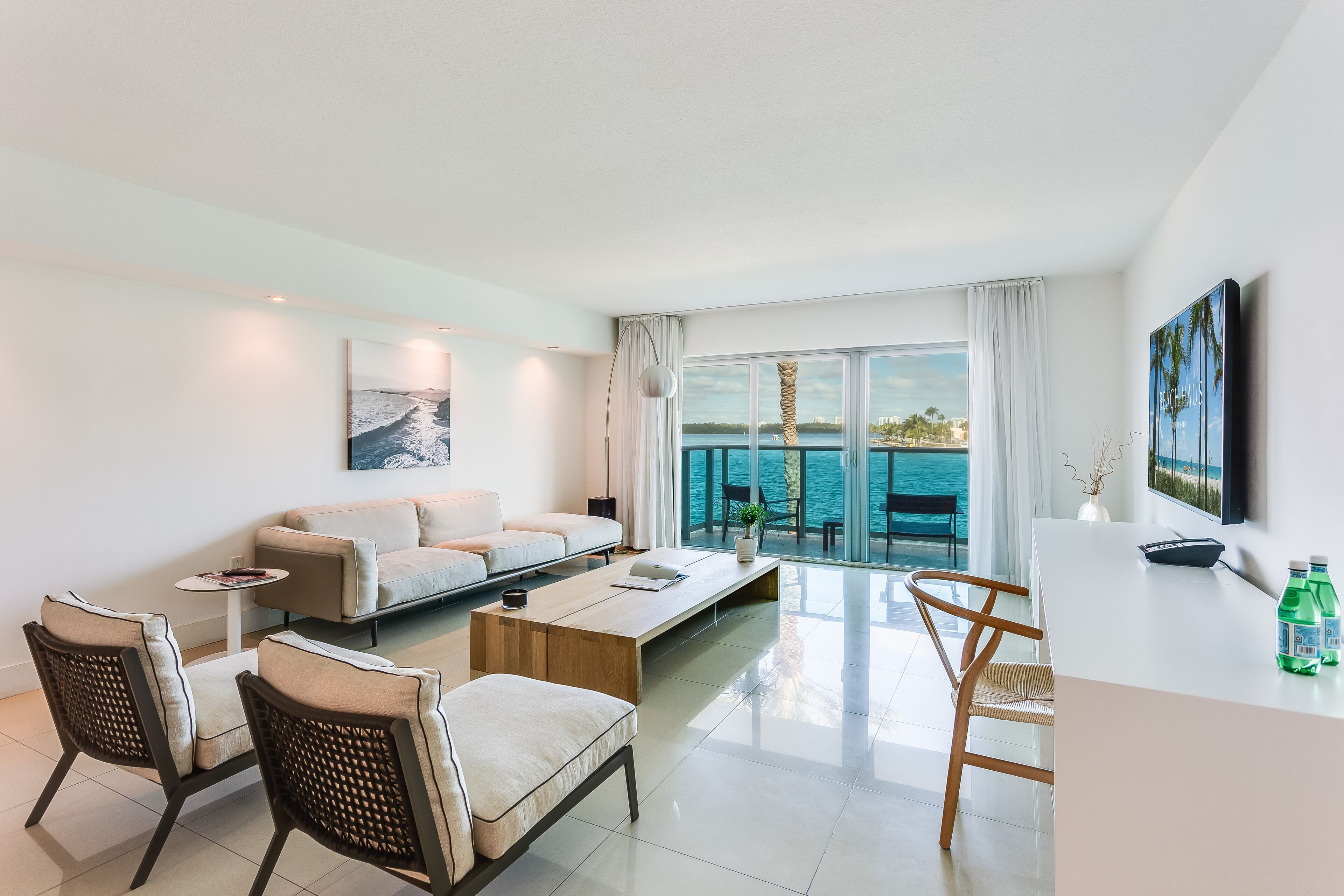 Un salon moderne comprenant un canapé, deux fauteuils, une table basse et un téléviseur. Une grande fenêtre offre une vue sur la mer.