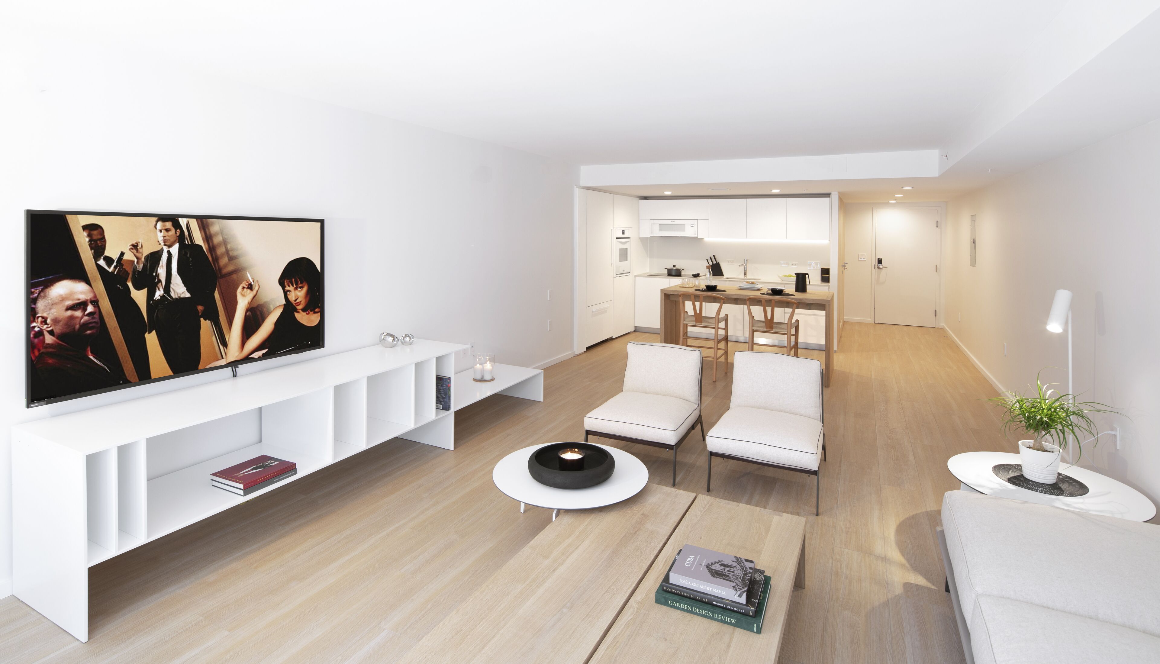 Una sala de estar moderna con un televisor de pantalla plana, sofás blancos, una mesa de centro y una cocina al fondo.