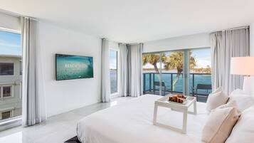 Una habitación de hotel moderna con una cama grande, una mesita con una bandeja de comida, un televisor montado en la pared y un balcón con vistas al mar.
