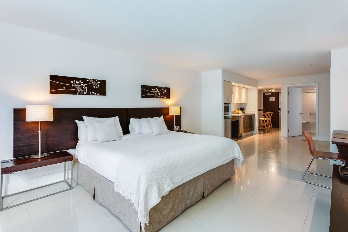 A modern hotel room with a large bed, bedside lamps, a headboard, and a view into the kitchen area.