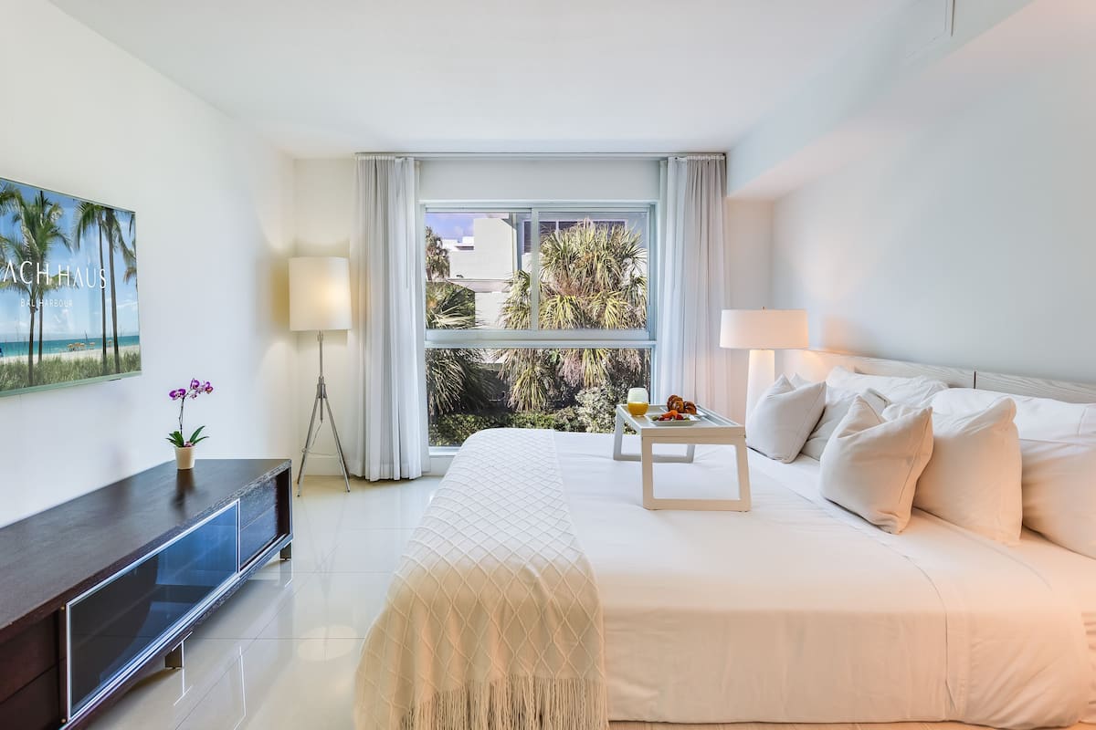 A hotel room with a large bed, a bedside table with a lamp, a small table with fruit, a TV, and a view of palm trees and buildings outside.