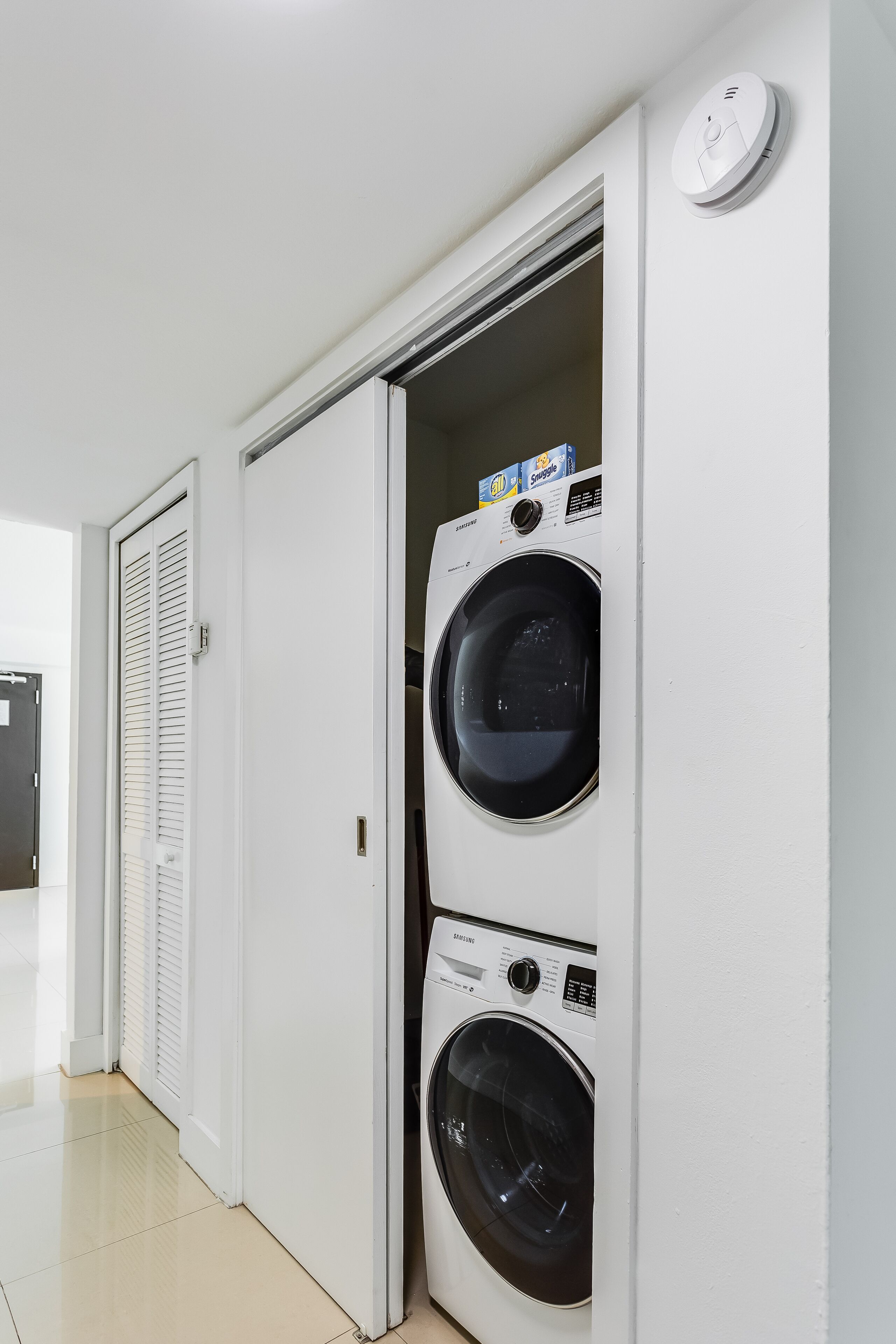 A laundry room with two stacked washing machines, a door, and a smoke detector on the wall.