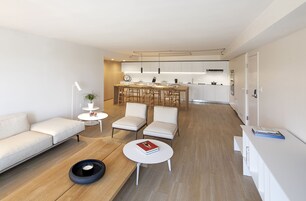 Uma sala de estar moderna com um sofá, duas poltronas, uma mesa de centro e um espaço de refeições, com uma cozinha ao fundo.