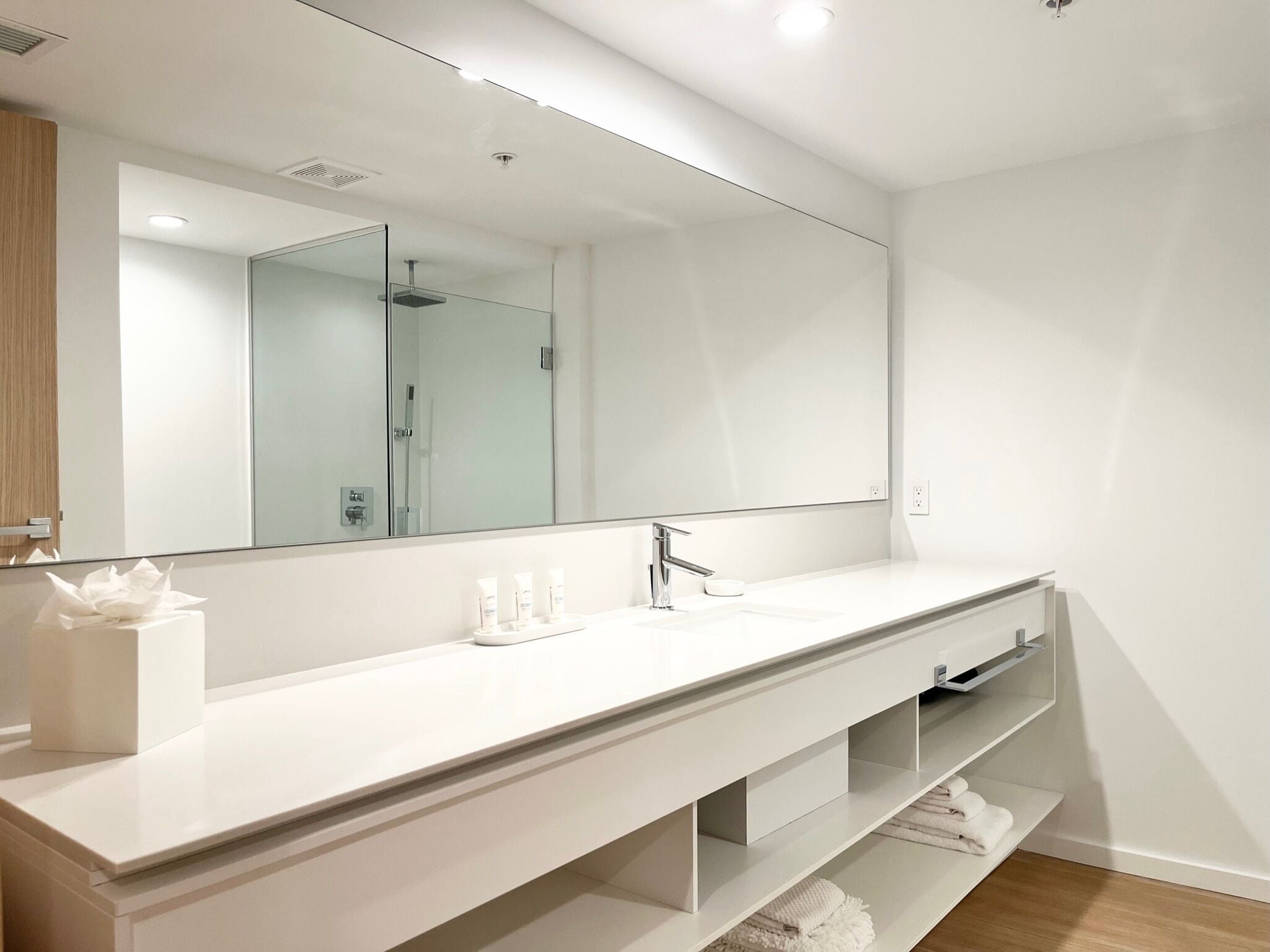 A modern bathroom with a large mirror, white vanity, and a shower visible in the reflection.