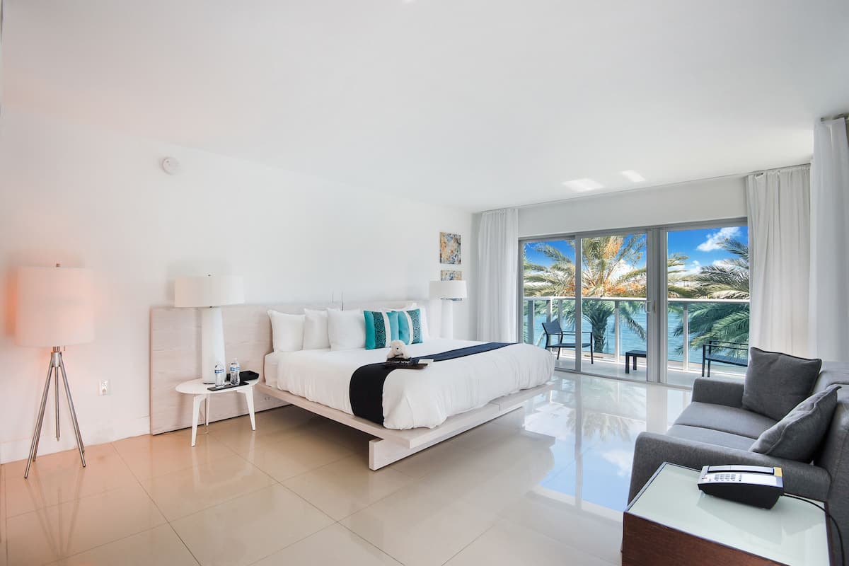 A modern hotel room with a bed, a sofa, a small table, and a large window offering a view of palm trees and the sea.