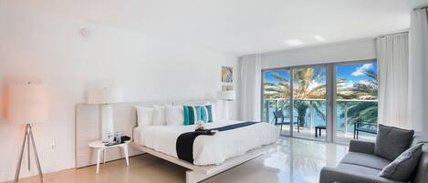 Una habitación de hotel moderna con cama, sofá, una mesita y un amplio ventanal con vistas a palmeras y al mar.