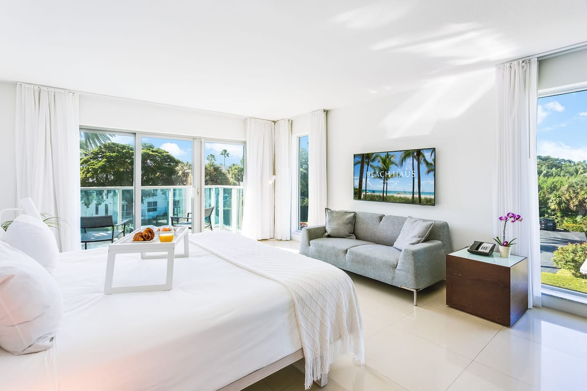 A modern hotel room with a large bed, a grey sofa, a small side table, and a balcony with a view of palm trees and a pool.