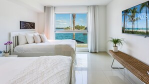A hotel room with a large window offering a view of the beach and ocean, two beds with white linens, a potted plant, and a framed picture on the wall. - Beach Haus Bal Harbour (Bal Harbour)
