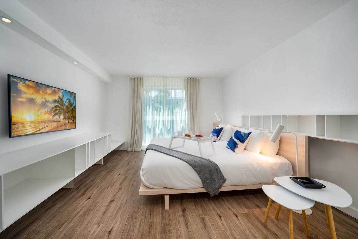 A modern hotel room with a wooden floor, a bed with white bedding and blue pillows, a bedside table with a lamp, and a large painting on the wall.