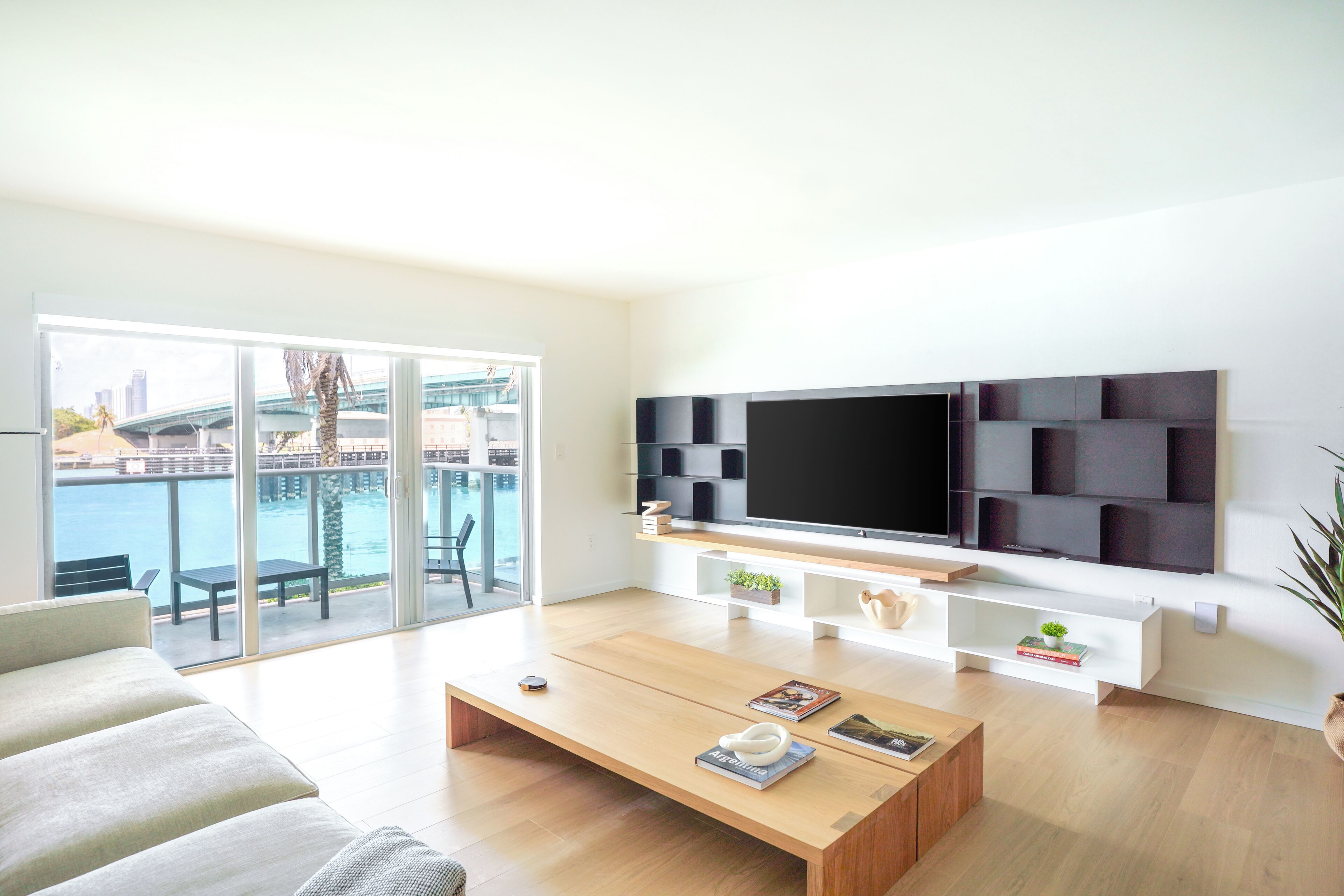 A modern living room with a large TV, a sofa, a coffee table, and a balcony with a view of the water.