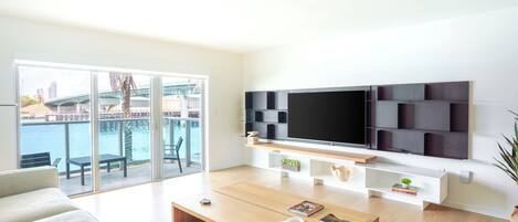 A modern living room with a large TV, a sofa, a coffee table, and a balcony with a view of the water.