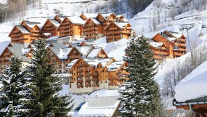 A cluster of wooden mountain lodges with multiple balconies, surrounded by snow-covered trees and a ski lift in the background.