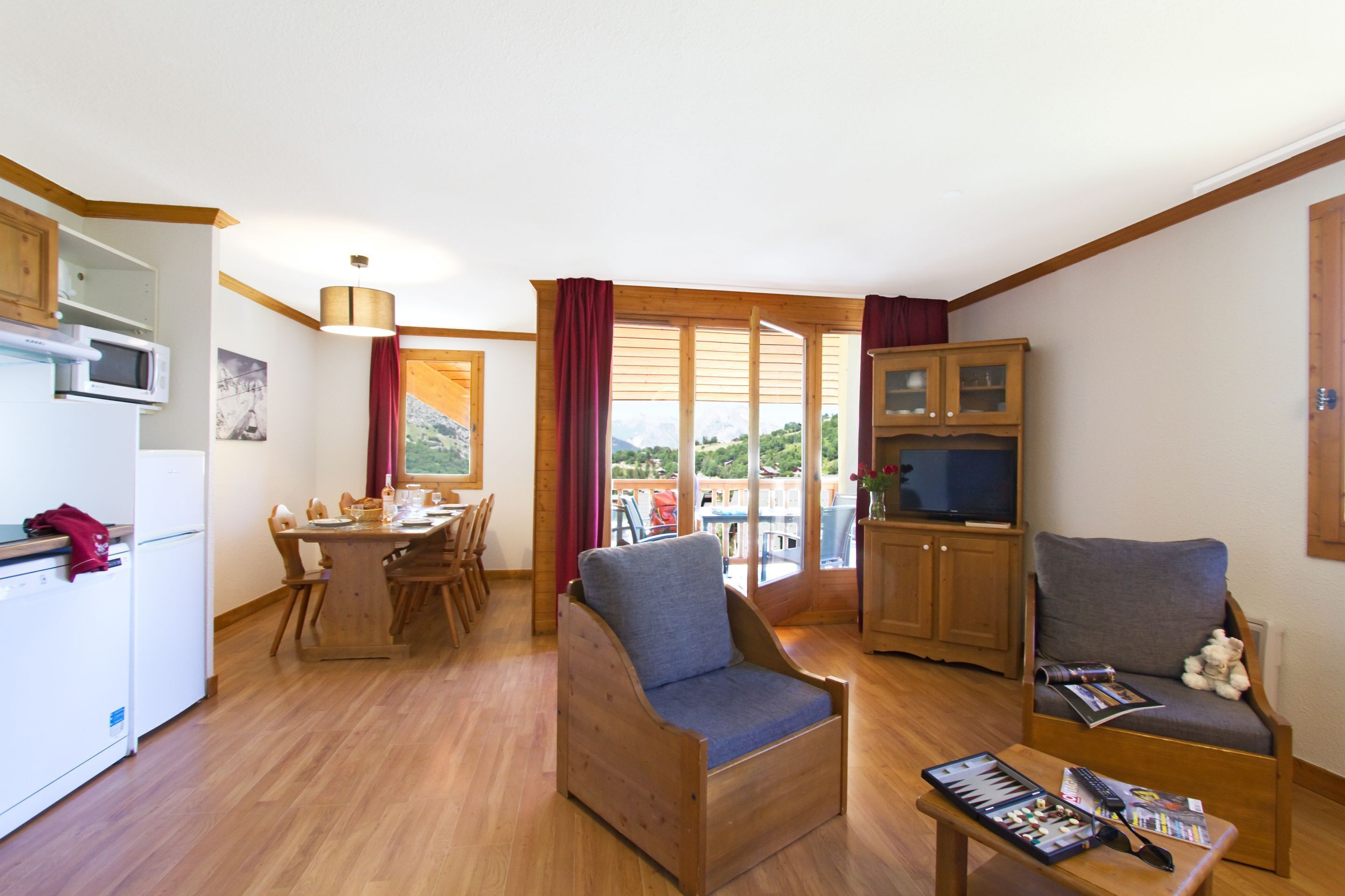 A compact living space with a dining area, two armchairs, a small table with magazines, a television, and a kitchen area with a microwave.