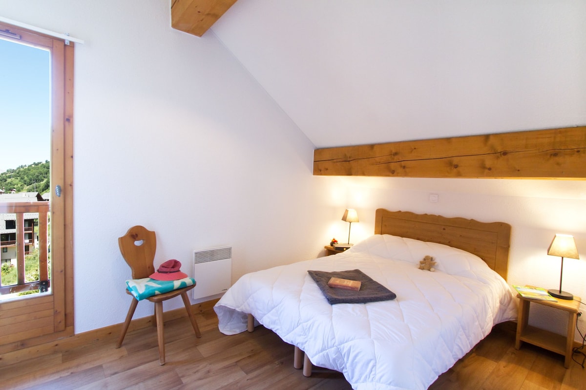 A bedroom with a wooden headboard, a single bed, a chair with a cushion, a bedside table with a lamp, and a window overlooking a balcony.