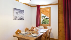 A dining area with a wooden table set for four, a window with a view of greenery, and a mountain landscape painting on the wall.