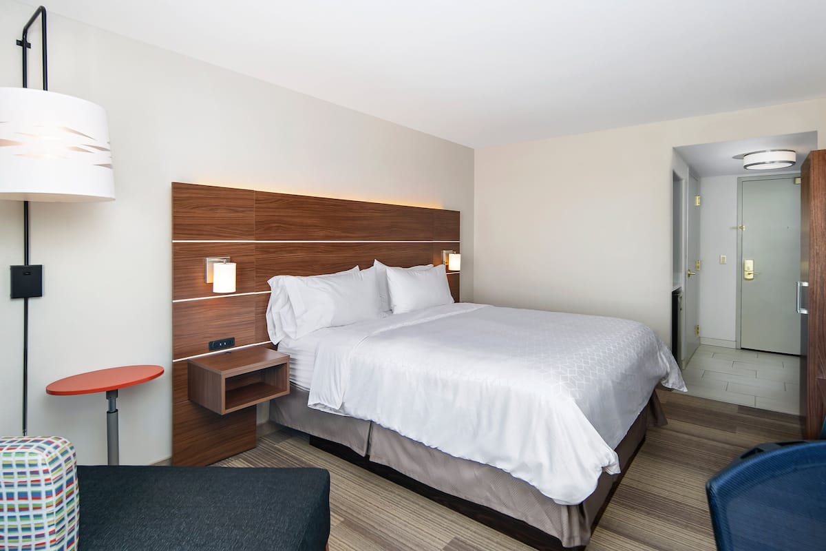 A hotel room with a large bed, a bedside table, a chair, a small table, and a wall-mounted lamp.