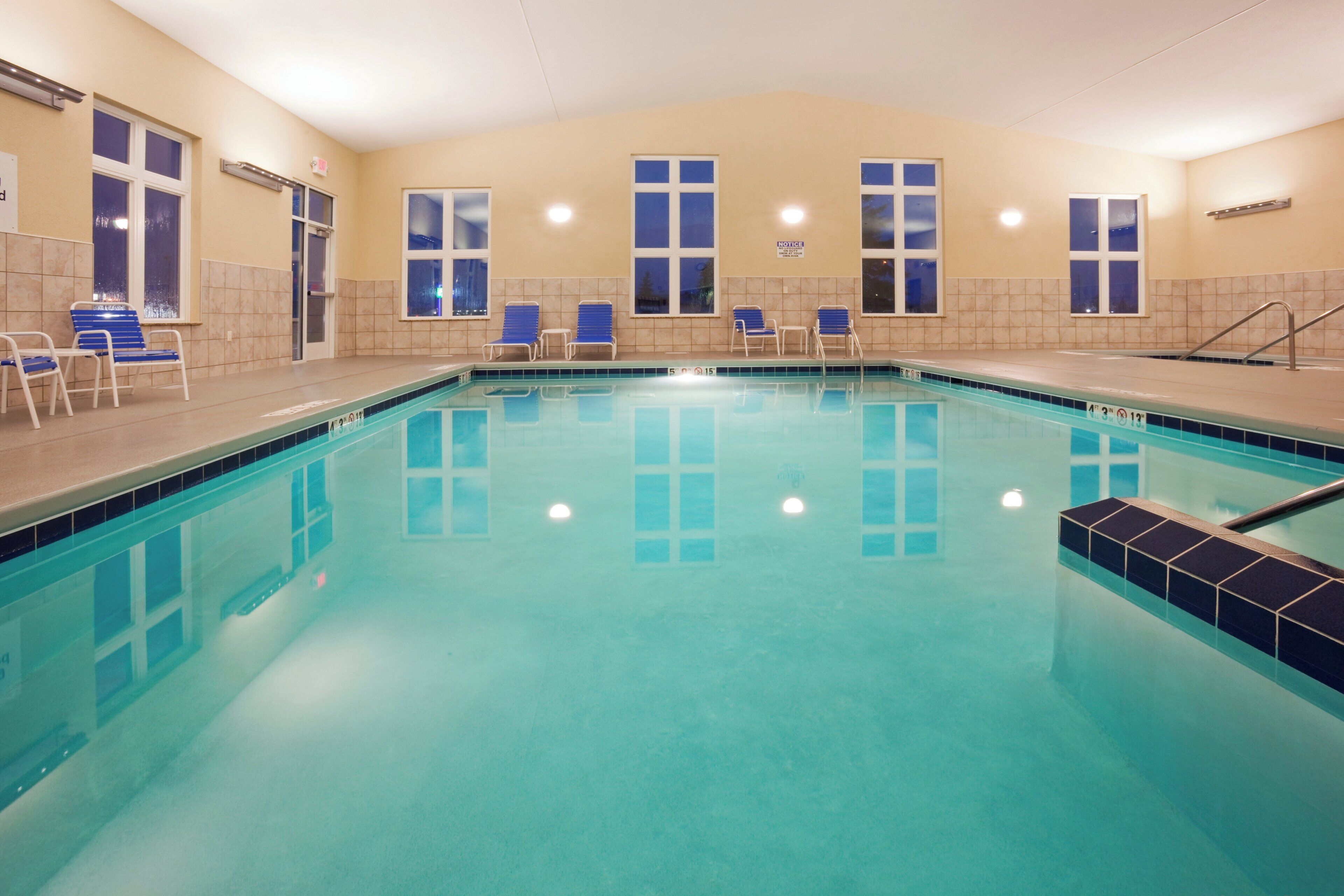 A swimming pool with clear blue water, surrounded by lounge chairs and a tiled wall.